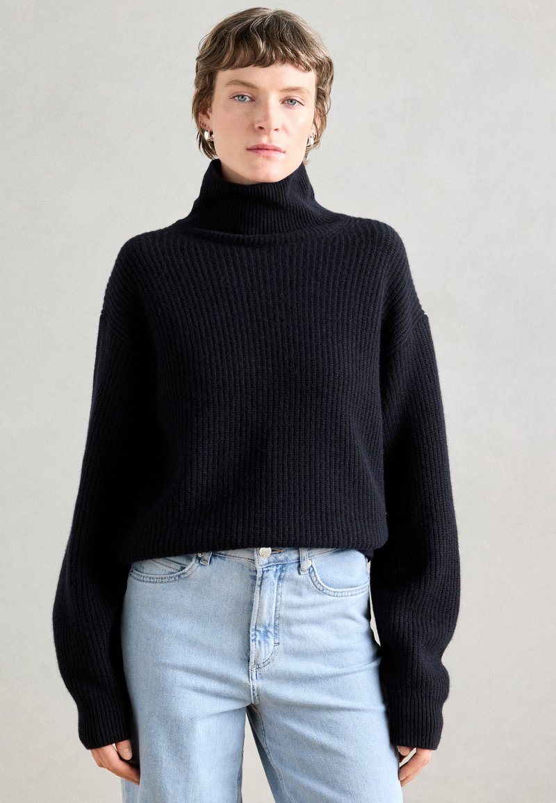 Μαύρο πλεκτό πουλόβερ με ριμπ μληστή λαιμόκοψη και χαλαρούς ώμους, συνδυασμένο με ανοιχτό μπλε τζιν. Μαλακή υφή και χαλαρή σιλουέτα.
