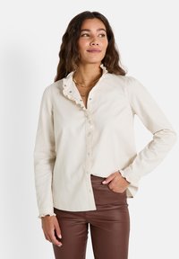 Blouse couleur crème avec un col volanté, des manches longues et un devant à boutons. Le tissu semble doux avec une légère texture. La coupe ajustée met en valeur la silhouette.