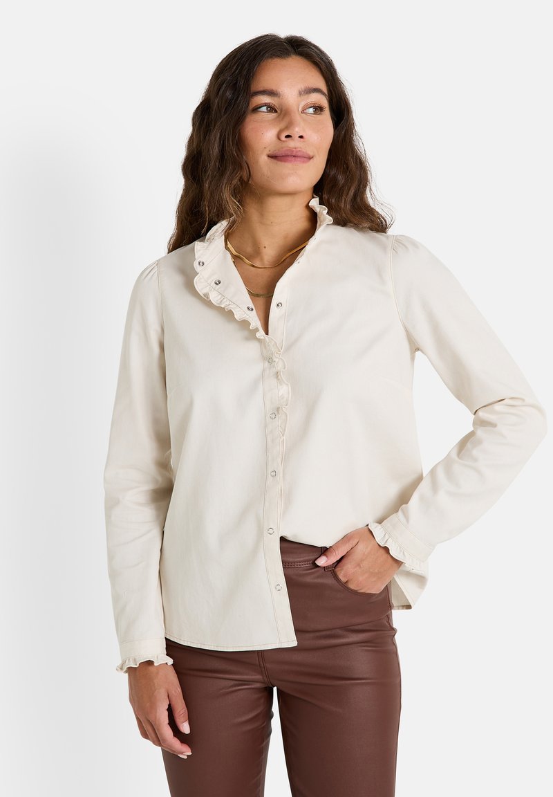 Blouse couleur crème avec un col volanté, des manches longues et un devant à boutons. Le tissu semble doux avec une légère texture. La coupe ajustée met en valeur la silhouette.