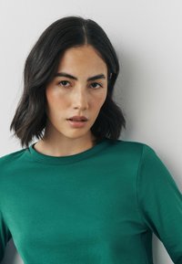 Grünes T-Shirt mit kurzen Ärmeln aus glattem, leichtem Stoff mit klassischem Rundhalsausschnitt. Besitzt eine lockere Passform und ein schlichtes, unauffälliges Design.