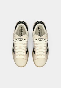 Beige suede sneakers with black stripes, featuring a white lace-up front and rubber sole. Adidas logo on the tongue and heel.