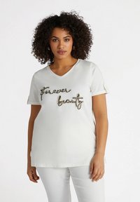 Camisa blanca de manga corta y escote en V con la frase "Belleza eterna" en adornos metálicos negros y dorados, con detalles de mangas enrolladas.