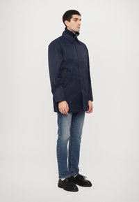 Navy blue, long coat with a tailored collar, button closures, and side pockets, paired with light blue jeans and black shoes.