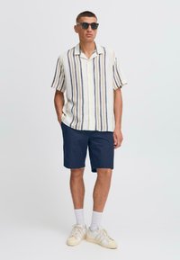 Chemise à manches courtes à rayures beige et bleues, assortie à un short bleu marine. Portée avec des lunettes de soleil et des baskets blanches avec des touches claires.