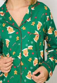 Groen pyjama bovenstuk met rode piping, voorzien van een patroon van peperkoeken huisjes, sneeuwvlokken, zuurstokken en peperkoeken mannen. Katoenen stof.