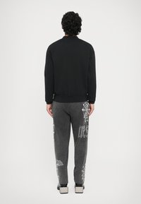 Sudadera negra con cuello redondo, combinada con pantalones deportivos grises que presentan gráficos de telarañas blancas y texto a lo largo de los laterales.