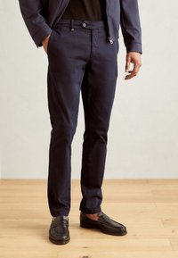 Pantalon en coton bleu marine avec une coupe slim, fermeture à boutons et poches latérales, associé à des mocassins noirs sur un fond de parquet en bois.