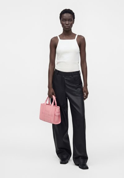 Femme portant un haut blanc sans manches et un pantalon noir à jambes larges, tenant un petit sac cabas rose avec des poignées.