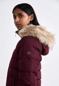 Bordo puffer jakna s kapuljačom od umjetnog krzna, vidljivo kviltana tekstura i okrugli logo na rukavu.