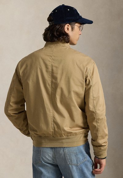 Keskmise pikkusega juustega mees, kes kannab prille, tumesinist nokamütsi, beeži jopet ja siniseid teksaseid, näidatuna tagant neutraalse tausta vastu.