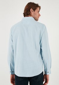 Camisa azul claro de manga larga en una tela texturada, con botones en los puños y un cuello clásico, mostrada desde la parte trasera.