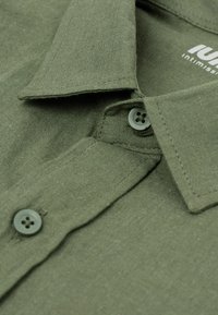 Olijfgroene shirt van een gestructureerde stof, met een puntige kraag en twee grijze knopen op de sluiting. Merkenlabel gedeeltelijk zichtbaar.