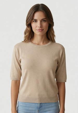 Mujer con cabello castaño ondulado hasta los hombros, vistiendo un suéter de manga corta beige y vaqueros azul claro, mirando hacia adelante sobre un fondo liso.