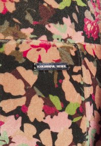 Látka s květinovým vzorem v odstínech růžové, zelené a hnědé. Obsahuje malou kovovou etiketu s nápisem "COLOURFUL REBEL." Texturovaný povrch.