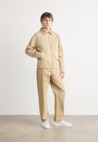 Beige denim jakk, millel on tõmblukk, kokku sobivad beežid püksid ja valged tossud. Lihtne disain, millel on krae ja küljetaskud.