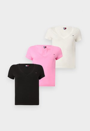 Γυναίκα με σγουρά σκούρα μαλλιά που φοράει μαύρο μπλουζάκι με λαιμόκοψη V, άνετο μπλε τζιν και λευκά αθλητικά, στέκεται μπροστά σε ένα απλό φωτεινό φόντο.