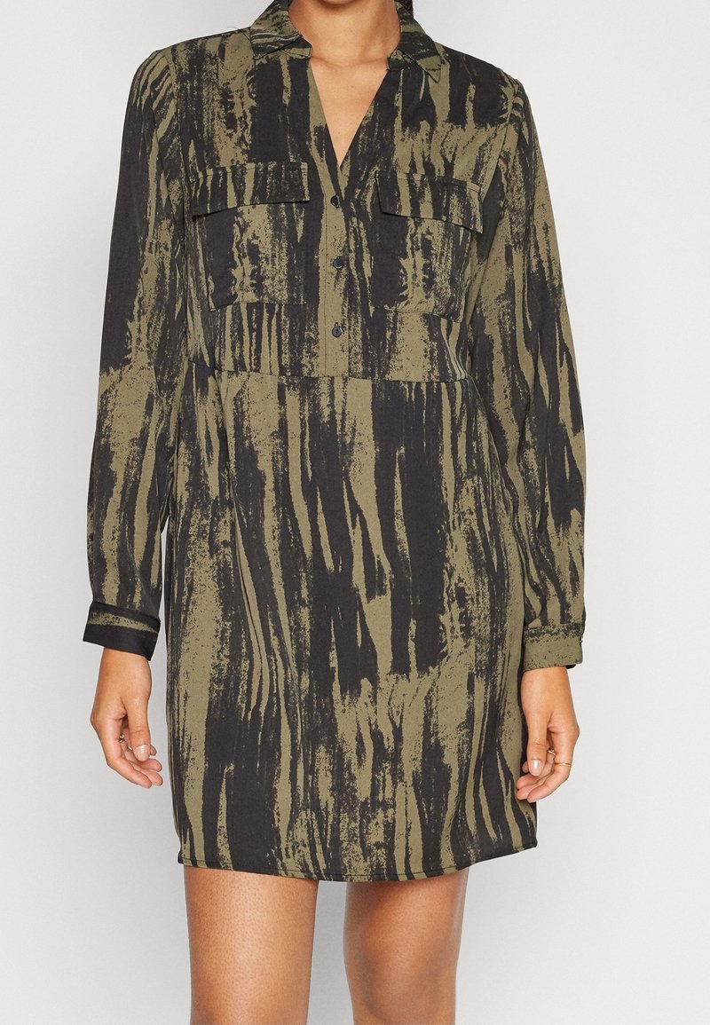 Robe à motif vert et noir avec un col, des manches longues et deux poches poitrine. Le tissu semble lisse avec une finition légèrement brillante.