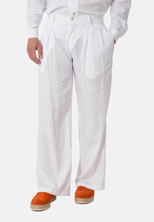 Uomo che indossa pantaloni bianchi di lino plisciati e larghi e scarpe slip-on arancioni in camoscio con suola di corda, in piedi con le mani in tasca.