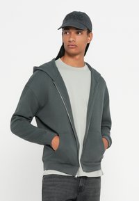 Felpa con zip di colore verde scuro, dotata di cappuccio con cordino, tasche frontali e polsini a coste. Indossata sopra una maglietta grigia chiara e abbinata a un cappellino grigio.