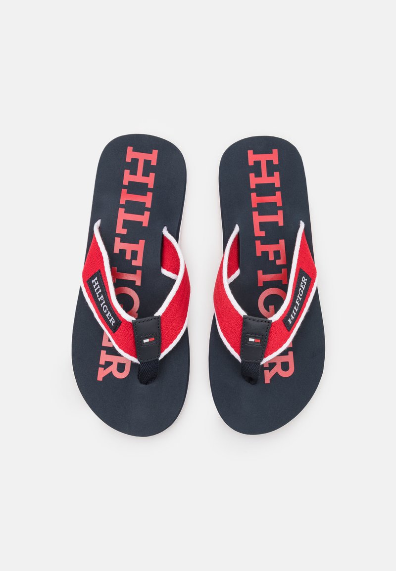 Sorte klipklapper med rød rem, der har hvide detaljer; stor rød "HILFIGER"-logo trykt på sålen, kanvasrem med logo-detalje.