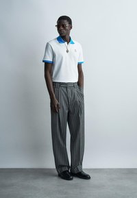 Bărbat purtând polo alb cu gulera și manșete albastre, pantaloni largi cu dungi, pantofi negri, ochelari de soare și colier cu pandantiv, stând în fața unui zid gri.