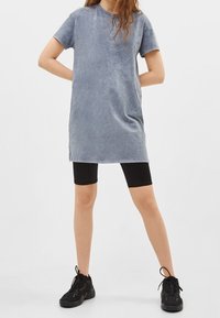 Szara sukienka krótko rękawowa o luźnym kroju i miękkiej fakturze, zestawiona z czarnymi szortami rowerowymi i czarnymi butami sportowymi.