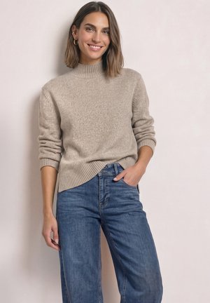 Femme souriante aux cheveux bruns mi-longs, portant un pull en tricot beige et un jean bleu, debout avec une main dans la poche devant un mur uni.
