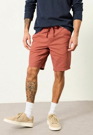 Man wearing rust-colored drawstring shorts, beige sneakers, white socks, and a dark blue long-sleeve shirt, standing on a gray floor.