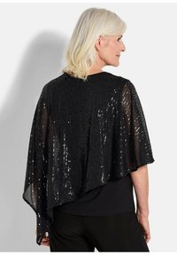 Schwarzer Pailletten-Cape mit offenem Design, fließender Silhouette und glänzender Textur. Über einem schwarzen Top getragen, fällt er hinten asymmetrisch ab.