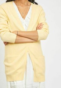 Light yellow cardigan with ribbed cuffs and hem, open front, and two front pockets, worn over a white dress with lace detail.