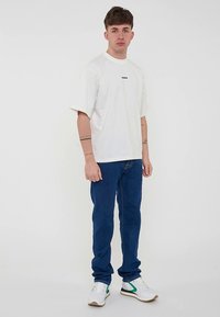 Ungdommelig mann som står og har på seg en hvit oversized t-skjorte, mørkeblå jeans, hvite sneakers med grønne detaljer, og tatoveringer på begge underarmene.