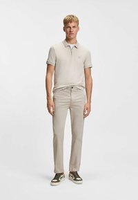 Beige Poloshirt mit Reißverschluss am Kragen, kombiniert mit beigen Hosen. Das Outfit zeichnet sich durch eine dezente Textur und Akzente von dunkelgrünen Sneakern aus.