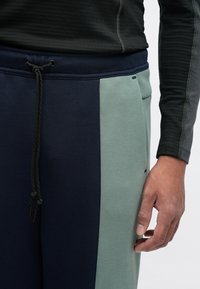 Φούτερ παντελόνια σε σχέδιο navy και πράσινου με μπλοκ χρωμάτων, διαθέτοντας ρύθμιση με κορδόνι στη μέση, μαλακό ύφασμα και πλαινές τσέπες. Κοντινή άποψη χεριών πάνω στο ύφασμα.