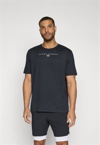 Schwarzes T-Shirt mit kurzen Ärmeln aus Baumwolle. Verfügt über einen Rundhalsausschnitt und das "UNDER ARMOUR"-Logo in Weiß, das über die Brust gedruckt ist.