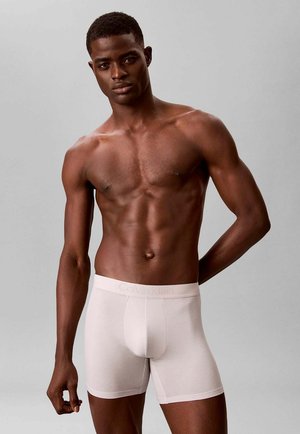 Muscular man with dark skin wearing light beige Calvin Klein boxer briefs, standing against a plain gray background.