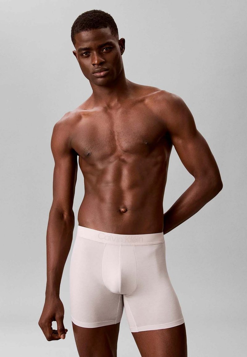 Gespierde man met een donkere huid die lichtbeige Calvin Klein boxershorts draagt, staand tegen een effen grijze achtergrond.