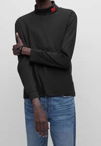 Schwarzes Langarm-Polohemd mit erhöhtem Kragen und rotem Hugo-Label, kombiniert mit blauen Jeans. Glatte Textur, lockere Passform.