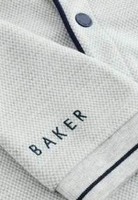 Hellgrauer Strickstoff mit einem strukturierten Muster, versehen mit einem navyblauen, gestickten Logo "BAKER" und einem kontrastierenden navyblauen Streifen am Saum.