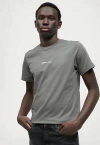 T-shirt de algodão cinza com mangas curtas e gola redonda. Apresenta um logotipo branco "REPLAY" na frente. Combinado com jeans escuros.