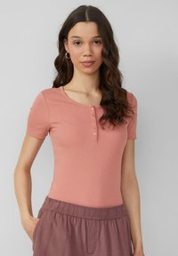 Różowa, prążkowana bluzka z krótkim rękawem o okrągłym dekolcie i pięcioma guzikami, zestawiona z luźnymi, fioletowymi spodniami na gumkę w talii.