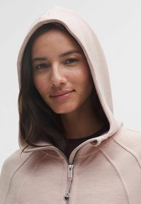 Hanorac roz deschis cu fermoar, dintr-un material texturat, având glugă cu șnur și detalii din metal argintiu la fermoar. Vedere detaliată a gâtului și feței.