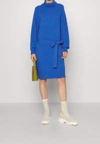 Abito blu lavorato a maglia con collo alto, vita cinturata e polsini e orlo a costine. Abbinato a stivaletti beige e una borsa verde.