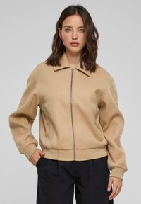 Beige zip-up jakke med krage, som har en avslappet passform, lange ermer og ribbestrikkede mansjetter. Mykt stoff med jevn tekstur.