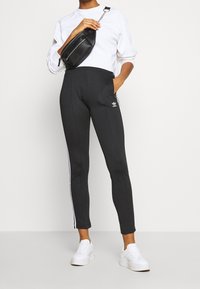 Černé slim-fit atletické kalhoty s bílými bočními pruhy, spárované s bílou mikinou a černou krosnou, doplněné bílými teniskami.