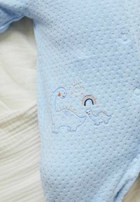 Monedero de algodón de color azul claro con tejido texturizado y patrones de puntos. Dinosaurios, sol y arcoíris bordados en colores suaves.