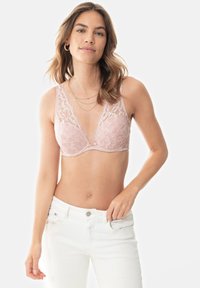 Krajková podprsenka v jemně růžové barvě s květinovým vzorem, se zavinovacím výstřihem a nastavovacími ramínky, v kombinaci s bílými džíny pro kontrast.