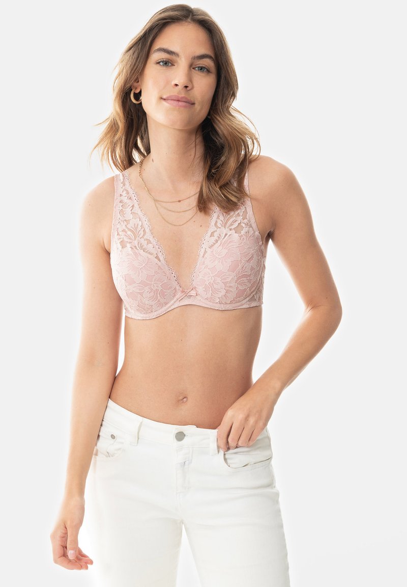 Krajková podprsenka v jemně růžové barvě s květinovým vzorem, se zavinovacím výstřihem a nastavovacími ramínky, v kombinaci s bílými džíny pro kontrast.