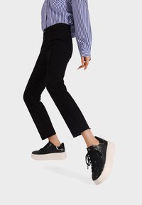 Persona che indossa pantaloni corti neri, sneakers platform nere con suola bianca e una camicia a righe blu e bianche, mentre compie un passo su uno sfondo bianco.