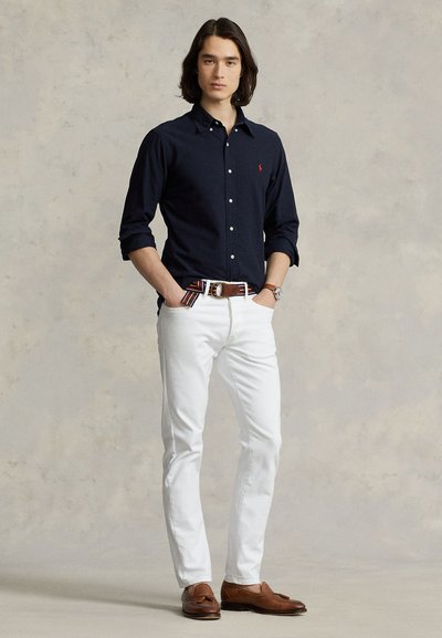 Uomo con capelli scuri fino alle spalle indossa una camicia blu navy, pantaloni bianchi, cintura a righe, mocassini marroni e un orologio, in piedi con le mani in tasca.