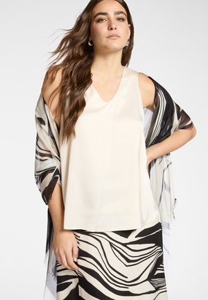 Beige mouwloos top met een V-hals en een zwart-wit patroon transparante sjaal over de schouders gedrapeerd; lichtgewicht stof, vloeiend ontwerp.